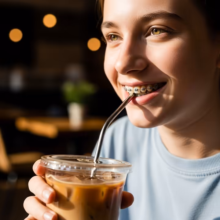 A smiling person with dental braces carefully drinking iced coffee through a straw to protect their teeth.