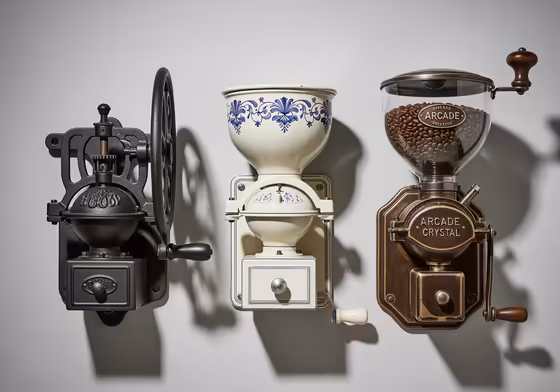 A lineup of three different styles of vintage wall-mounted coffee grinders: a black cast-iron model, a ceramic Dutch model with blue paint, and an American Arcade model with a glass hopper.