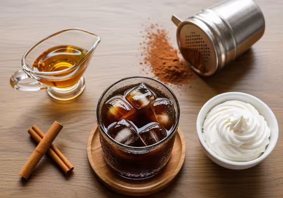 An assortment of items to flavor cold brew, such as a bottle of vanilla syrup, cinnamon sticks, a pitcher of milk, and a bowl of cold foam.