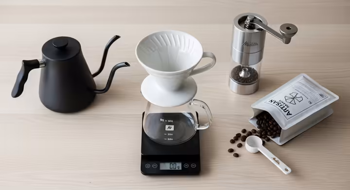 A flat lay photograph of all the essential gear for making Melitta pour over coffee: a ceramic Melitta brewer with paper filters, a gooseneck kettle, a digital scale, a timer, and a bag of whole bean coffee next to a burr grinder.