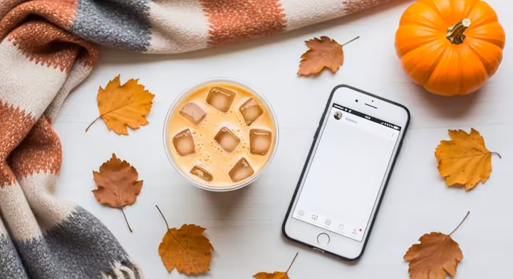 A top-down photo of an iced pumpkin spice latte on a wooden table, next to a smartphone displaying social media, sunglasses, and autumn leaves.