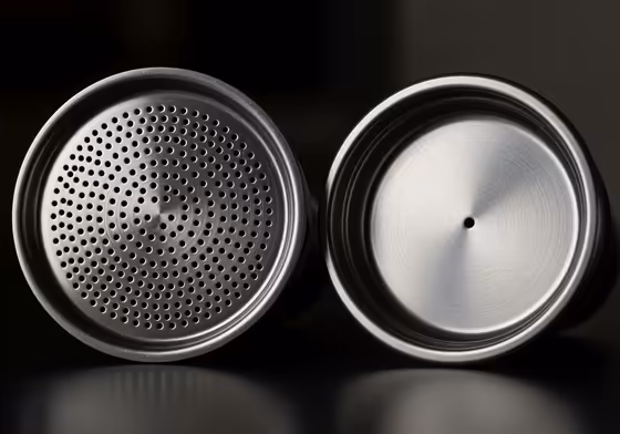 A close-up view comparing a non-pressurized espresso basket with hundreds of tiny holes to a pressurized basket with only a single exit hole.